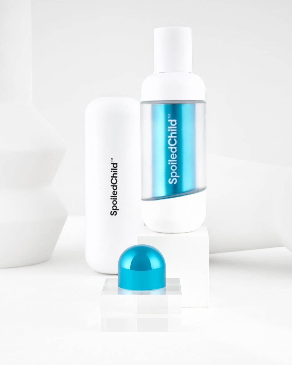 A product arrangement featuring a tall, white SpoiledChild S33 bottle with a teal capsule interior, alongside a white product box and a small teal cap, all set against a clean white background.
