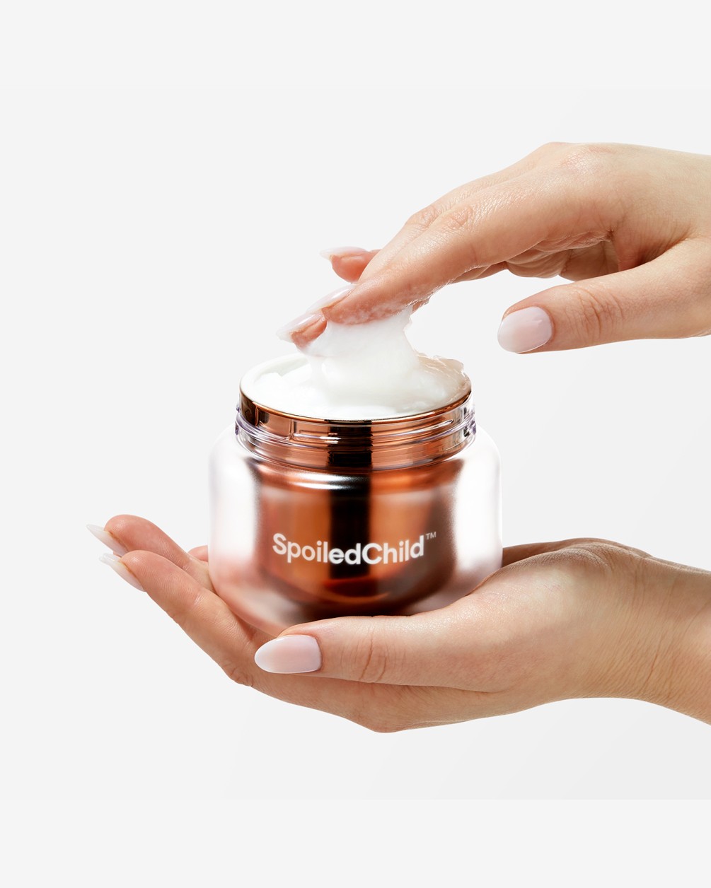 A close-up of a pair of hands holding an open, metallic copper-colored jar of SpoiledChild M26 hair mask. One finger is scooping a thick, white cream from the jar, highlighting its rich, buttery texture against a minimalist white background.