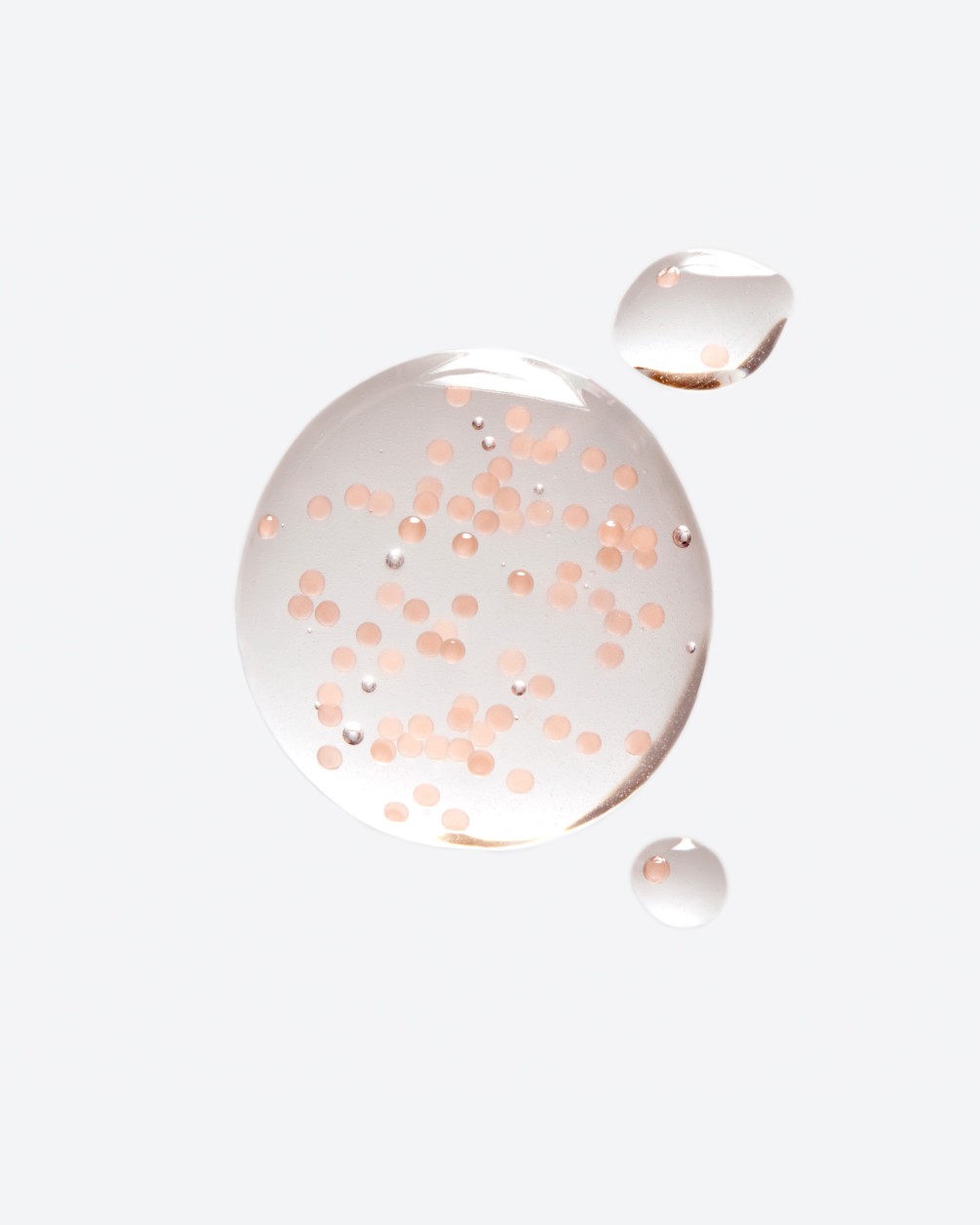 A close-up of several translucent, circular droplets of a liquid product on a white surface. The largest droplet contains numerous small, suspended pinkish-orange spheres or microbeads.