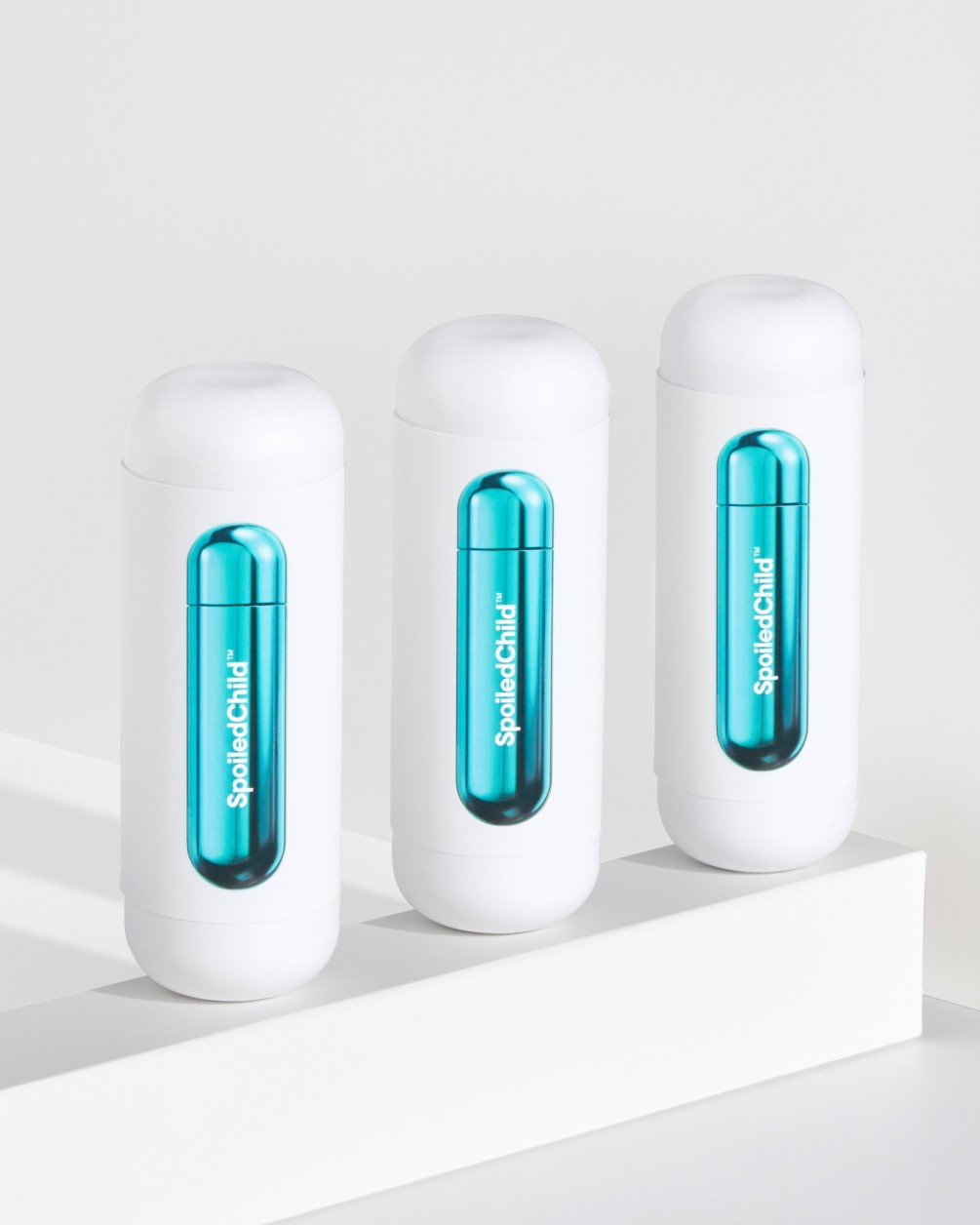 A product arrangement featuring three white SpoiledChild S33 containers lined up on a white pedestal. Each container has a window revealing a sleek, metallic teal capsule inside.
